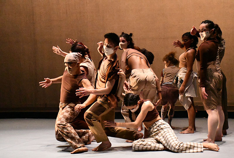 A group of dancers in earth-toned clothing and sepia-toned lighting oerform a modern dance involving a series of poses at varying levels 