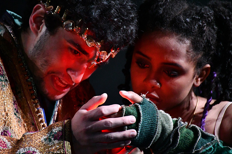 Closeup of an actor wearing a crown and an actor with layers of sleeves and fingerless gloves who are both looking at a glowing red light eminating from their hands