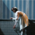 A young woman in a white lacy dresshunches over in dim lighting while running her fingers along the molding of a blue-and-white striped wall