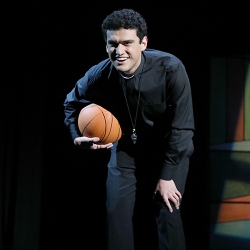 An actor with short brown wavy hair, dressed as a catholic priest in black with a white collar, leans over while holding a basketball