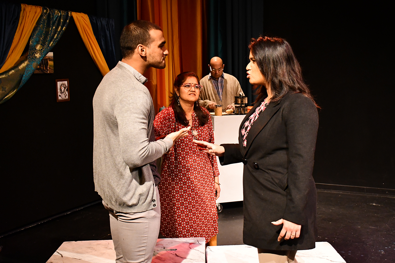 Two young adults face off in the foreground as a concerned older woman with a tika mark on her forhead looks on. In the background an older manwearing glasses appears to be preoccupied with a different task.