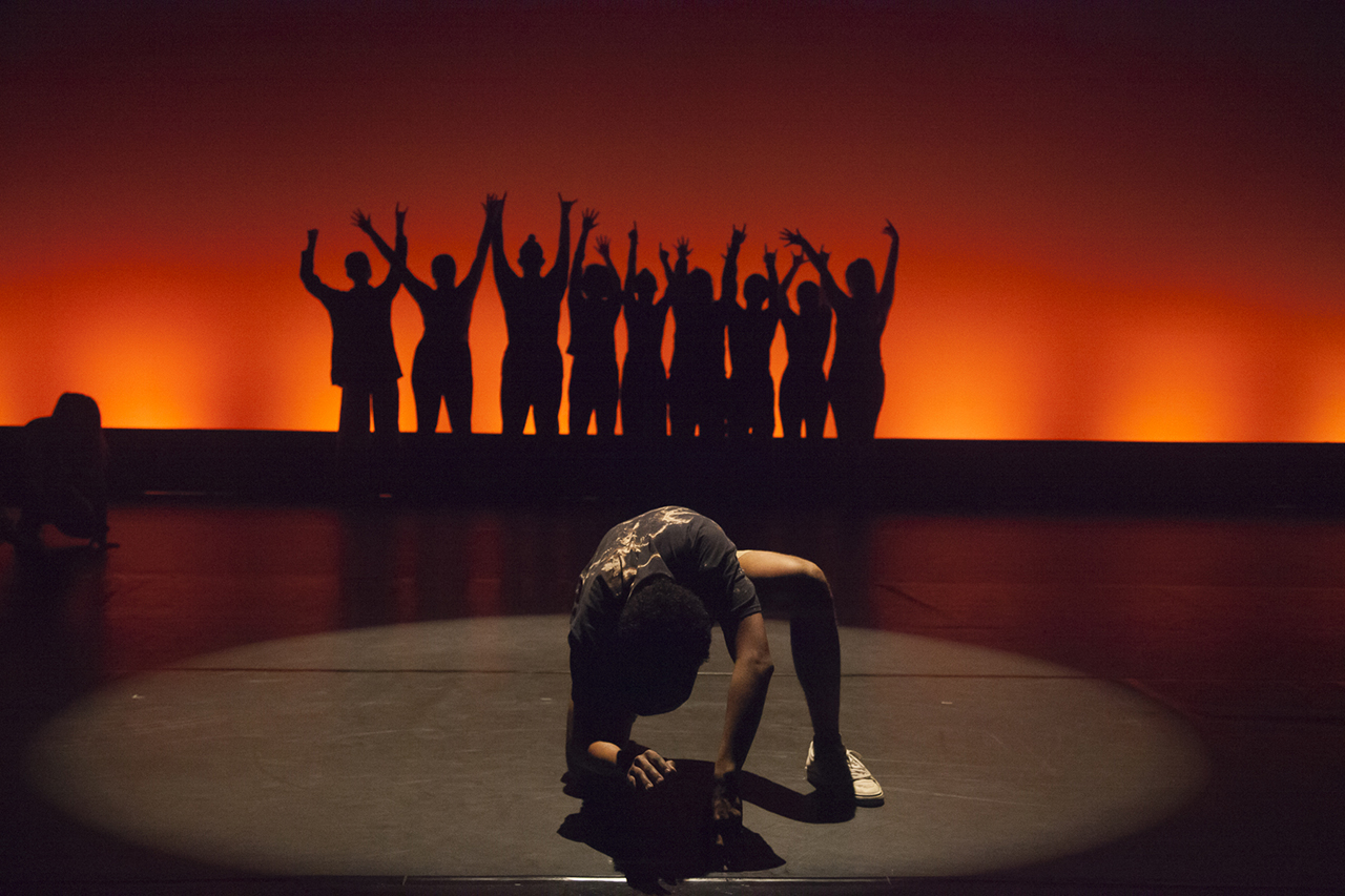 A stage backlit with a deep red to orange gradient against which a group of students are posing in silouette with their hands raised. A single dancer kneels on one knee in the foreground in a pool of light.