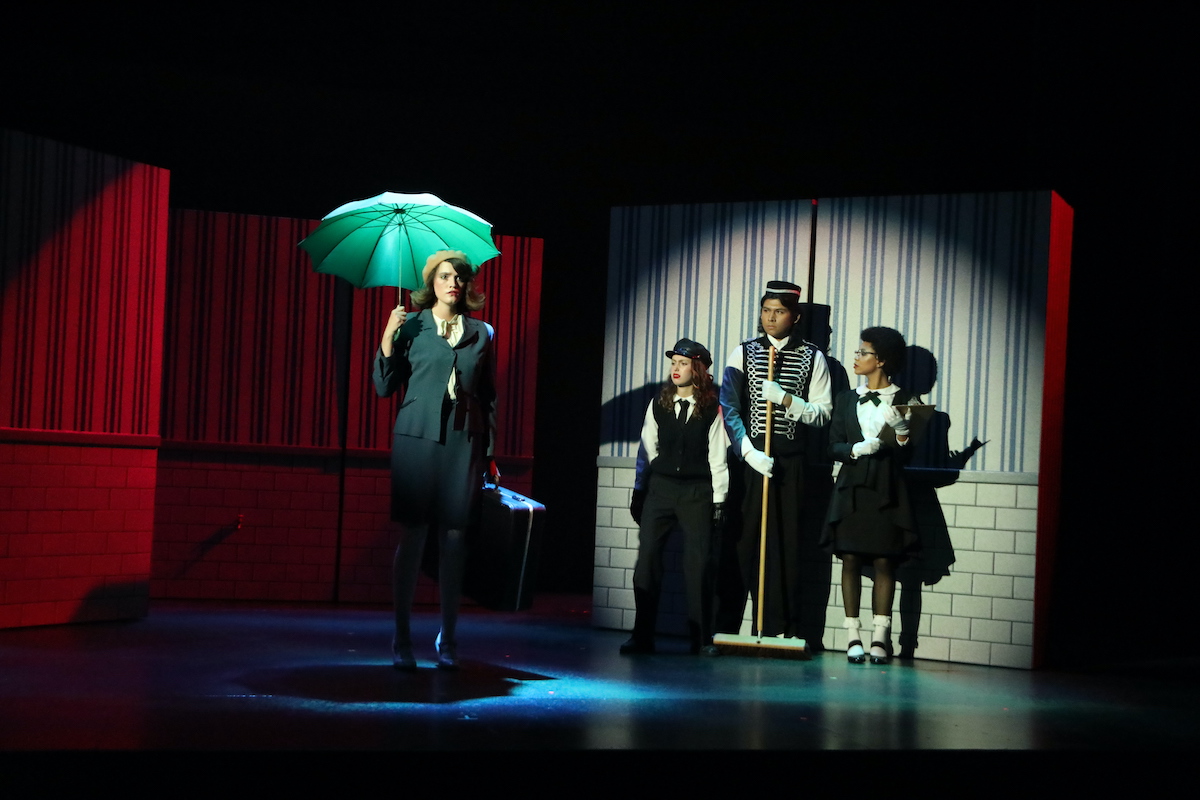 A woman in a blue suit and cap holding an open umbrella enters a spotlight watched by three actors dressed in bellhop suits against a striped wall.