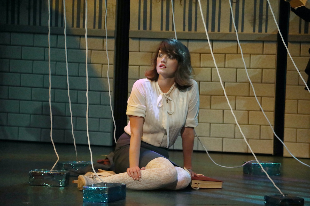 A woman in a white bluse and beret sits on a stage floor surrounded by pieces of string descending from above.