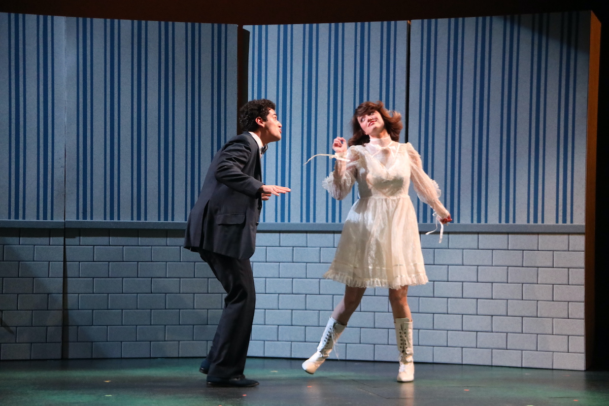 Two actors playing lovers are dancing, the male actor wears a dark suit, and the woman wears a white lace dress.
