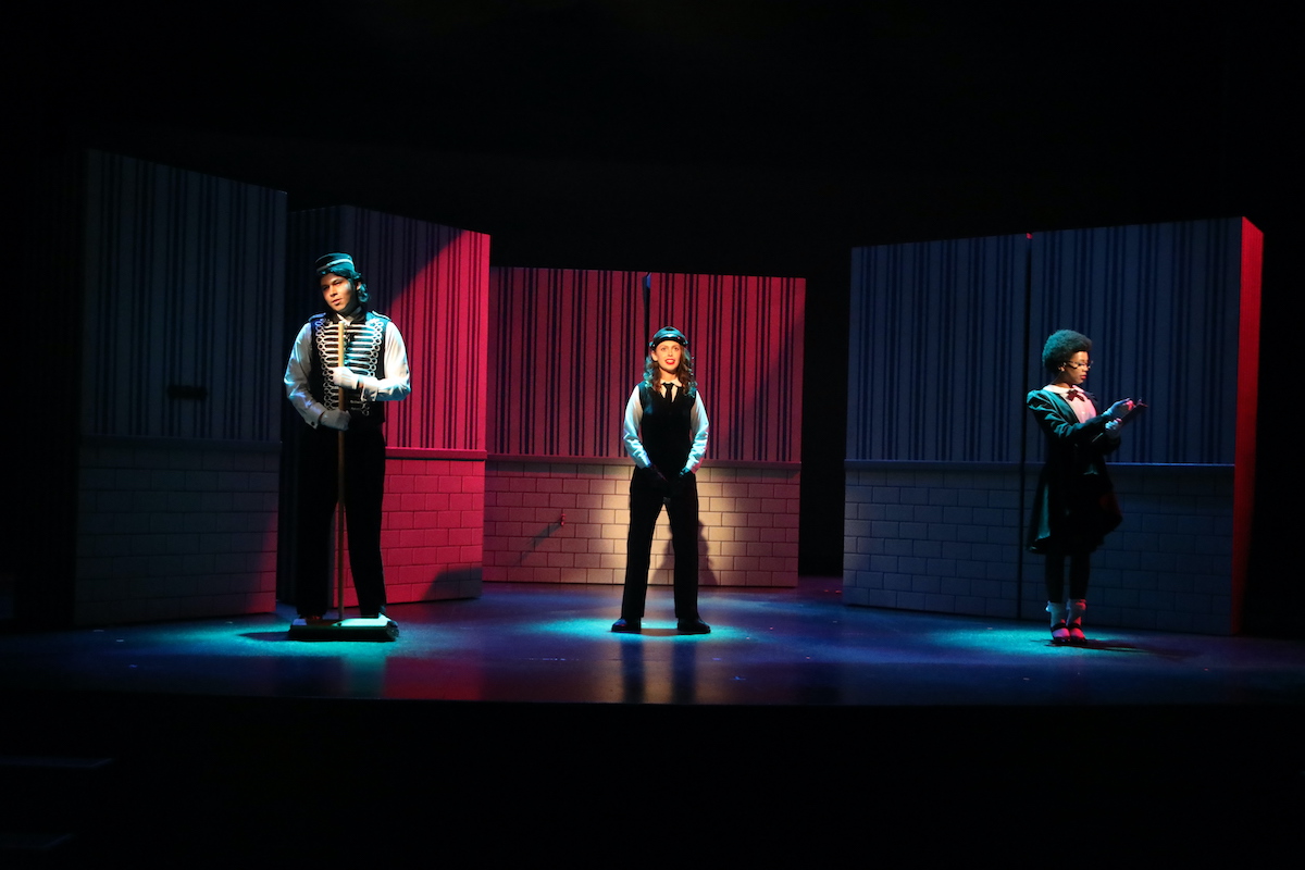 Three actors dressed in bellhop suits are standing in three spotlights against a striped wall.