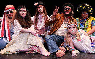 A group of student actors dressed in "hippie" costumes sitting together on a bare stage, throwing peace signs