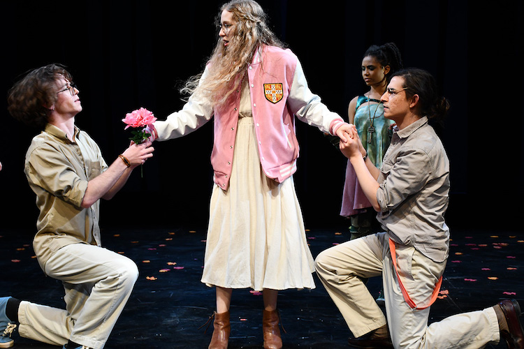 Two actors on bended knee flank a third actor with long blond hair and glases