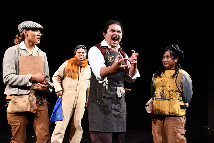 Group of four actors in workperson clothing, one appears to be roaring at the camera