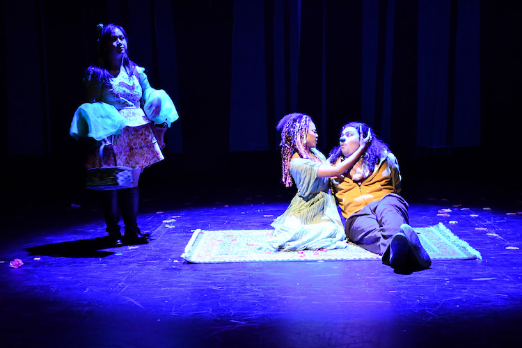 Two actors cuddle on a blanket in a purple light while a thrid actor approaches them from stage right
