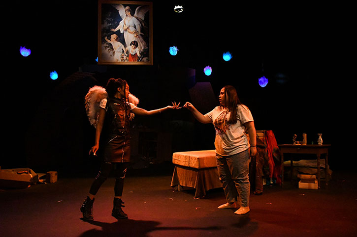 A Black actor dressed as an angel hands something to a Latinx actor in a Virgen of Guadalupe t-shirt. The lighting is dim and amber with glowing blue lights in the background and a painting of an angel with children