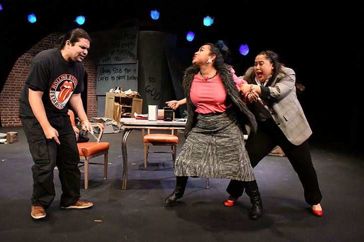 Three Latinx actors in an onstage fight. A tall male actor with a ponytail and Rolling Stones T-shirt, a female actor in a pink blouse, grey skirt, and black boots, held back by a female actor in a grey plaid jacket, black pants, and red shoes