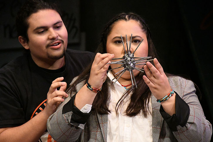 An actor in the role of "Lenny" shows off a metal wire sculpture to an actor playing "Marisol," telling Marisol it is a portrait of her