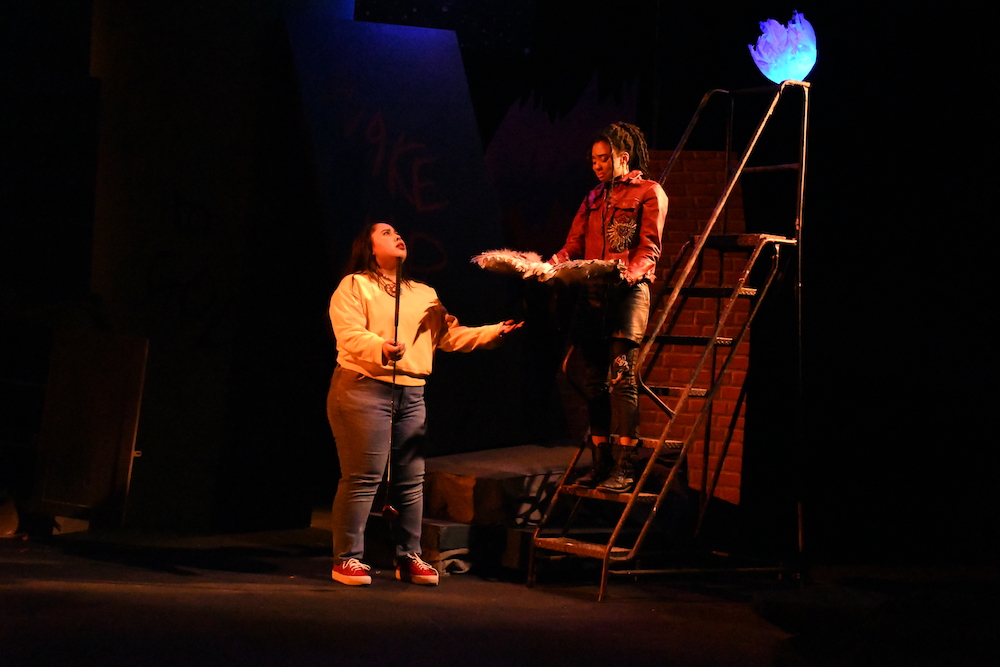 Actor playing Marisol, with long brown hair and a yellow sweatshort, confronts actor playing the angel standing on a stepladder with dredlocked hair and leather pants, holding her wings in her hands