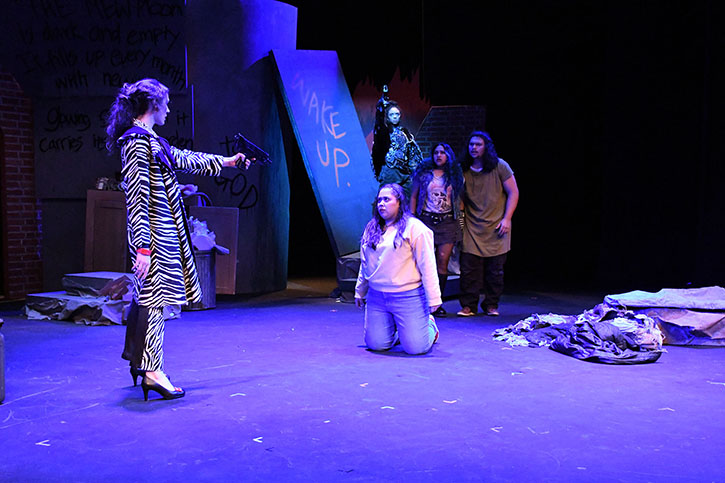 The actor playing the woman in furs dressed in zebra stripes holds a gun at the actor playing Marisol who is on her knees in blue light