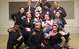 A group of actors in a cluster, dressed in all black and wering a variety of stylized half-masks made of silicone and other materials