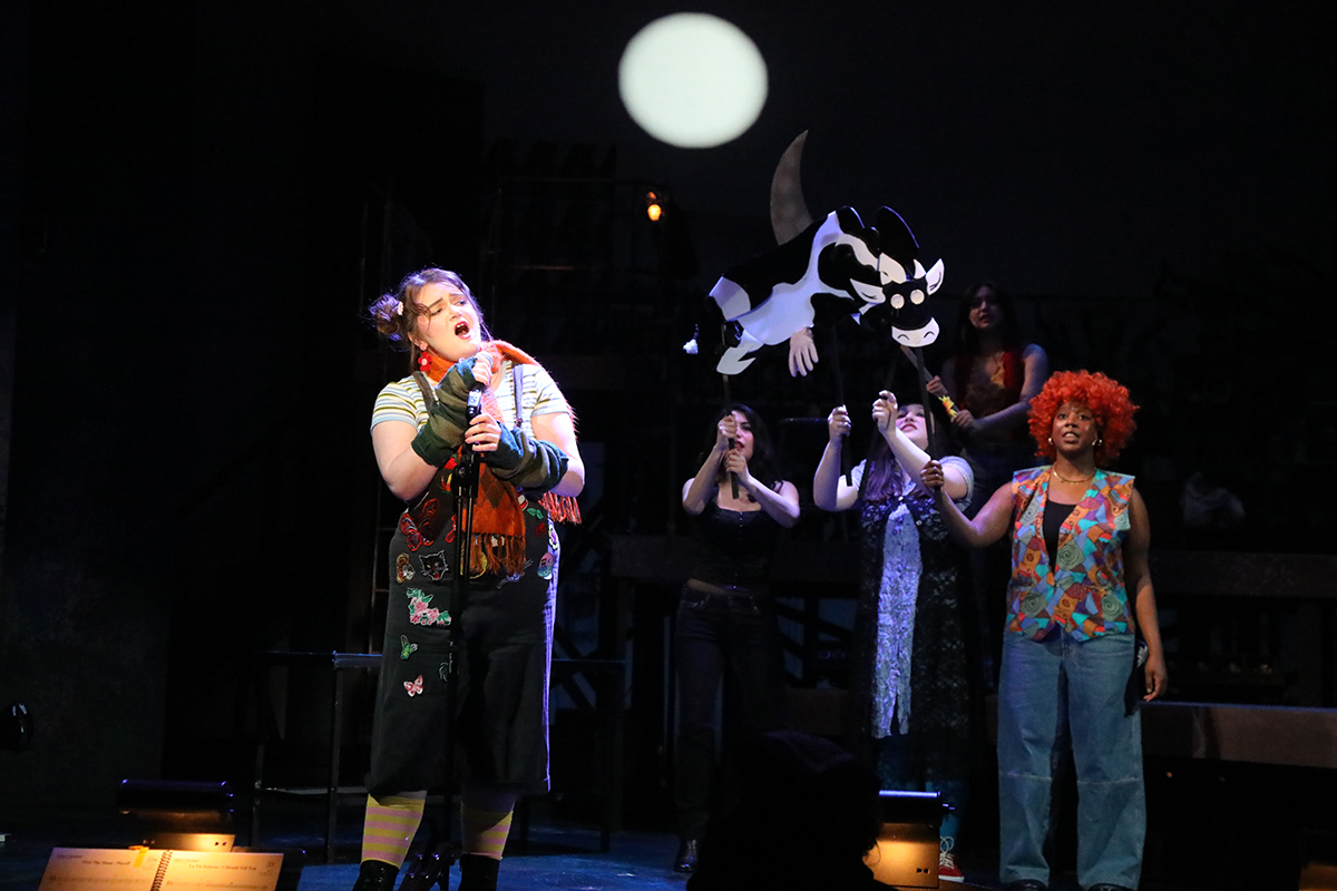 Actor dressed in embroidered overalls sings into a microphone as a cow puppet and a spotlight moon inhabit the background.