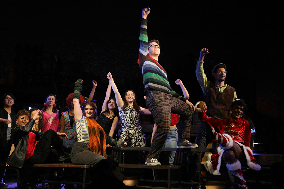 An ensemble of cast members of RENT in bohemian winter clothing such as striped seaters sit around a long table with their fists raised in the air. The actor playing the role of Mark stands on the table with his fist up, he is wearing glasses and a striped sweter of many colors.