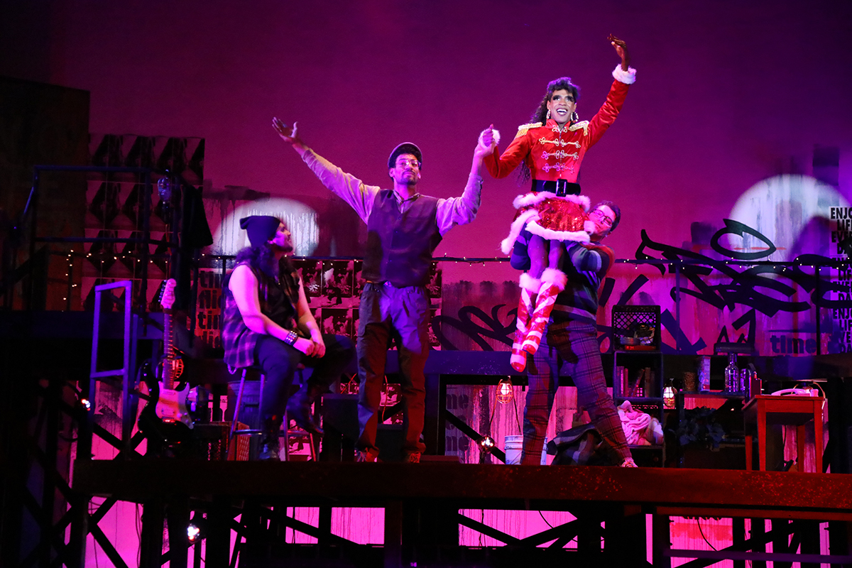 Four actors in 80s shabby chic dance onstage, one of whom is dressed in santa drag with a short red minishirt and white trim being held up in the air with their hand raised. The lighting is a saturated magenta color.