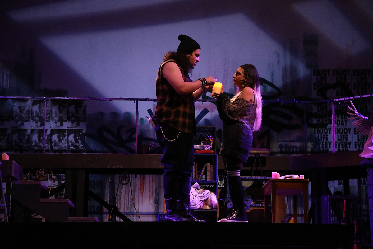 Two actors face each other, on dressed as a rock and roller in a cut off sleeveless flannel and a beanie, one dressed in a grey mini sweater dress and black knees socks with white stripes hold a lit candle between them and look into each other's faces.