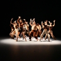 A group of dancers in earth-toned clothing and a pool of light pose in a circle with their hands in the air