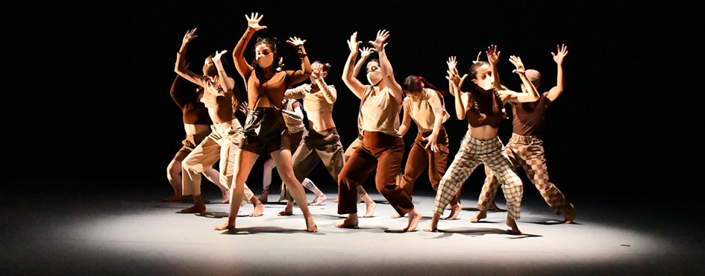 A group of dancers in earth-toned clothing and a pool of light pose in a circle with their hands in the air