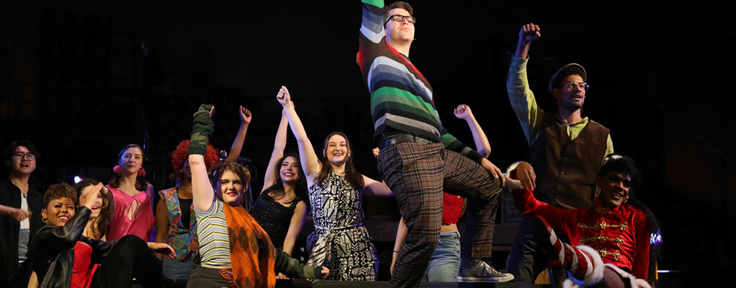 An ensemble of cast members of RENT in bohemian winter clothing such as striped seaters sit around a long table with their fists raised in the air. The actor playing the role of Mark stands on the table with his fist up, he is wearing glasses and a striped sweter of many colors.