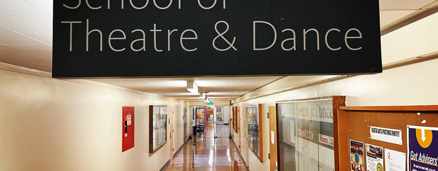 School of Theatre & Dance sign hanging above the hallway of the School. There are bulletin boards, doors, and lights stretching into the distance