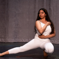 Mirian Hernandez action shot. A dancer with long dark hair, wearing white leggings and tank top crouches low to the floor with one leg extended out and their arms folded across their body.