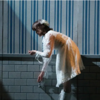A young woman in a white lacy dresshunches over in dim lighting while running her fingers along the molding of a blue-and-white striped wall