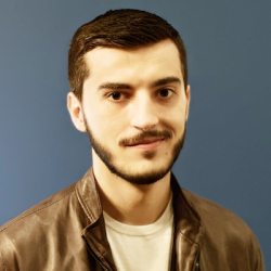 A younger man with short dark hair and a small trimmed beard and mustache wearing a brown leather jacket and t-shirt