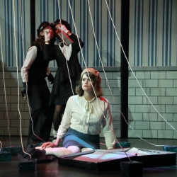 A white woman in a beret and white blouse sits on a stage surrounded by strands of string, two actors in bellhop suits stand behind her against a striped wall.