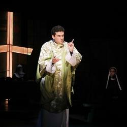 A cross of light shines behind an actor dressed in the robes of a Catholic priest who appears to be giving a serious sermon