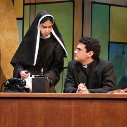 A young woman with a darker complexion and dressed like a nun stands over a young man with wavy dark hair dressed as a priest seated at a large wooden desk