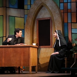 A young man with dark wavy hair, dressed as a priest and seated behind a large wooden desk smiles at a young woman dressed as a nun who is sitting on a chair to the left