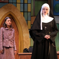 A young woman with a darker complexion wearing a smart-looking mauve suit from the 60s stares at the back of an actor dressed as a nun looking sternly into the camera