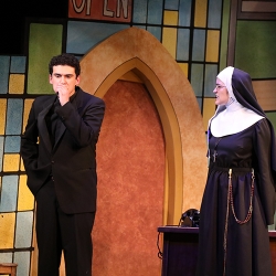 An actor with dark wavy hair dressed as a priest bites his hand nervously as an actor dressed as a nun looks at him and speaks to him angrily