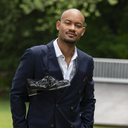 Black man standing with hands in pockets and tap shoes strung over right shoulder
