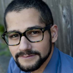 Ayounger man with dark, close-cropped hair, dark glasses, and a styled beard and mustasche stands against a tree and looks directly at the camera