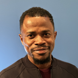 A Black man with close-cropped dark hair and a beard wears a dark brown jacket and a red t-shirt, smiling at the camera