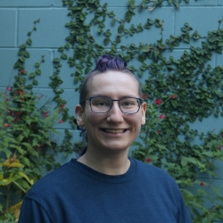 person with purple faux-hawk, and blue t-shirt, smiling widely in front of a teal painted wall that has ivy growing on it