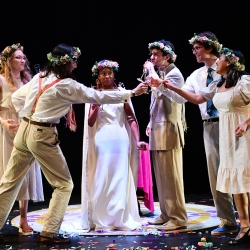 A group of six actors in festive wedding attire toast each other in a circle, confetti at their feet
