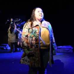 A Latina Actor in a patterned coat stands in a spotlight, as actors behind her lift an actor dressed as an angel into the air