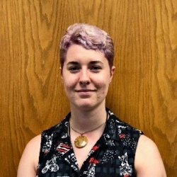 Person with short pink hair and patterned sleeveless blouse standing in front of wood paneling
