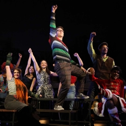 Ensemble cast of Rent sitting around a long table with their fists raised in the air. One actor wearing a striped sweater and glasses is standing on the table as if leading the chant.