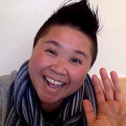 Headshot for Thao P Nguyen, an Asian woman with short, dark spiked hair smiing at the camera with her left hand raised as if in greeting
