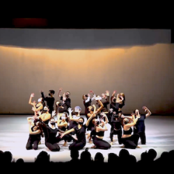 An ensemble of dancers in black dance clothing and black facemaskspose onstage at the end of a number