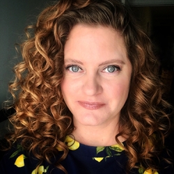 Vicki Hoskins, a white woman with curly reddish hair, looks at the camera. She is wearing a dark top patterned with bright yello lemons.