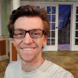 Carson Blickenstaff, a white man with short brown hair combed back and glasses smiles at the camera wearing a white sweatshort