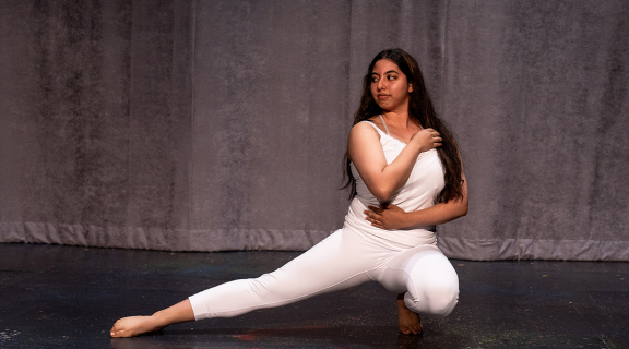 Mirian Hernandez action shot. A dancer with long dark hair, wearing white leggings and tank top crouches low to the floor with one leg extended out and their arms folded across their body.